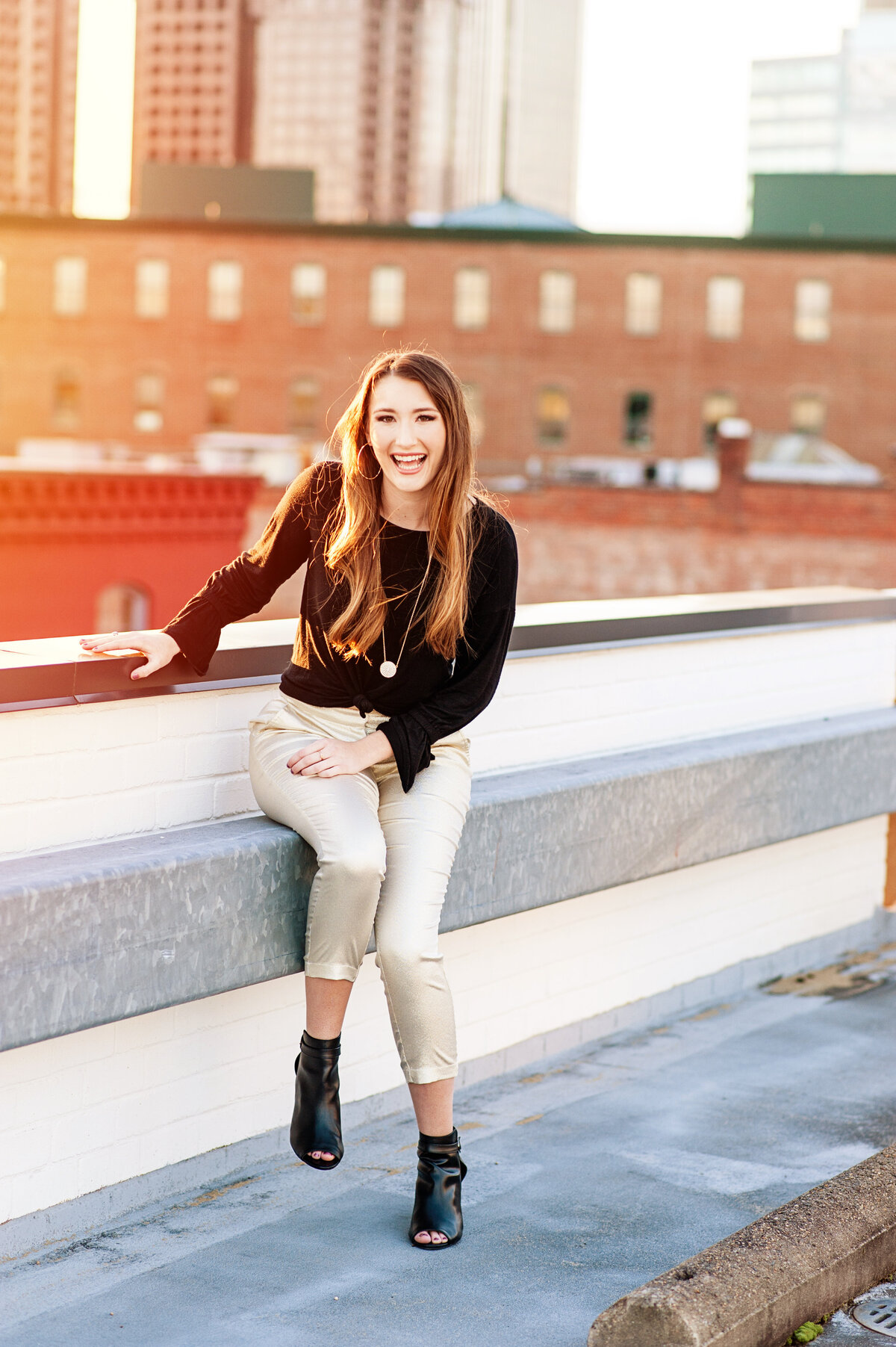 RVA senior girl poses on rooftop during her senior portrait session at sunset wearing gold pants.