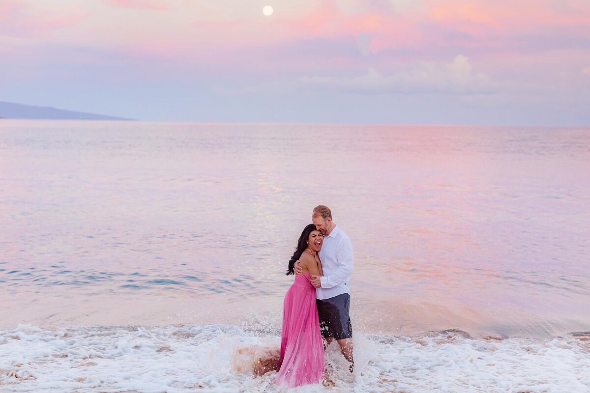 Couple kissing on the shoreline on Maui during their sunrise full moon session. Woman wears a pink gown and waves splash on them as they smile and laugh.