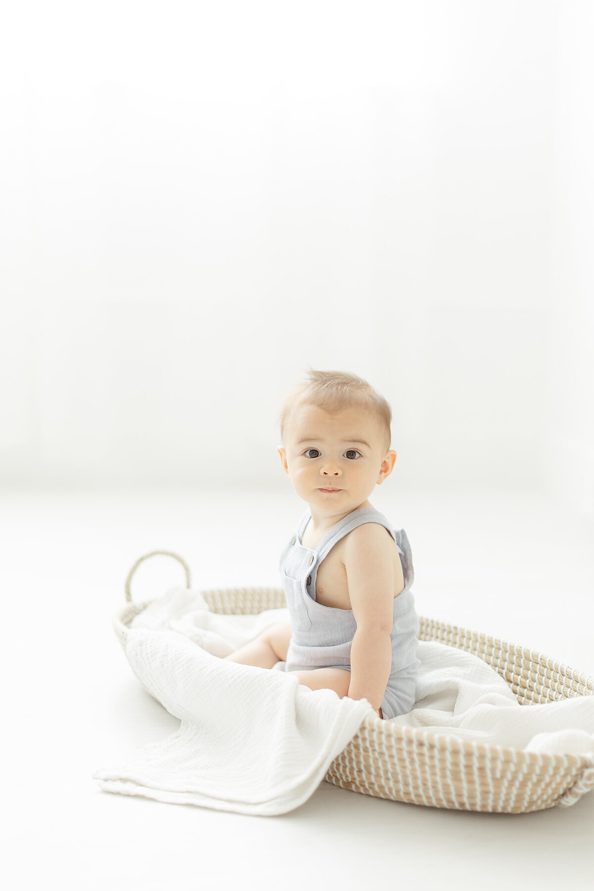 A cute little baby boy sitting in a basket in a photography studio by a window.