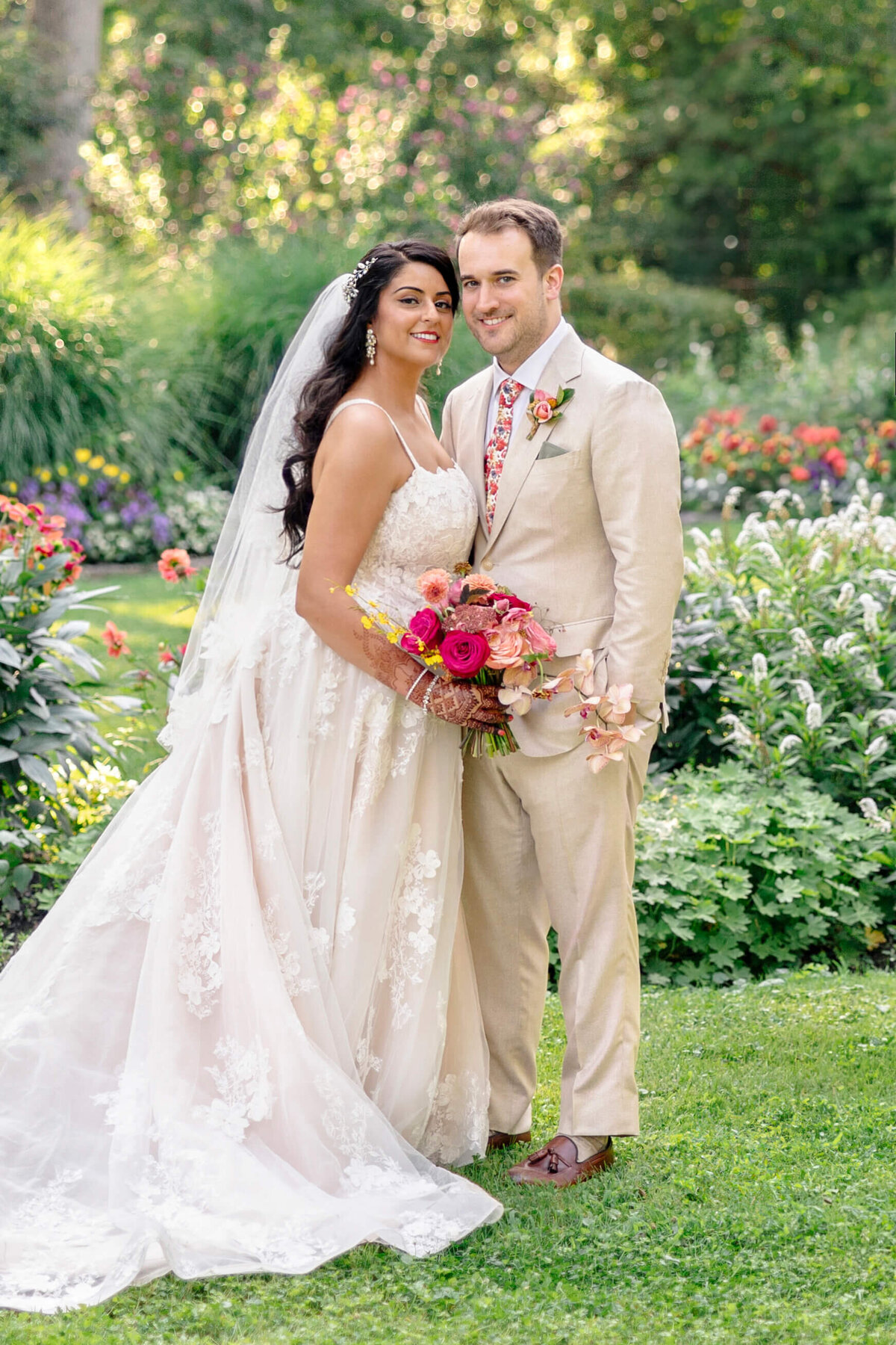 A bride and groom stand together in a lush garden. The bride wears a lace wedding dress and holds a bouquet of vibrant flowers, while the groom is in a beige suit with a floral tie. They are smiling, surrounded by green foliage and colorful flowers.