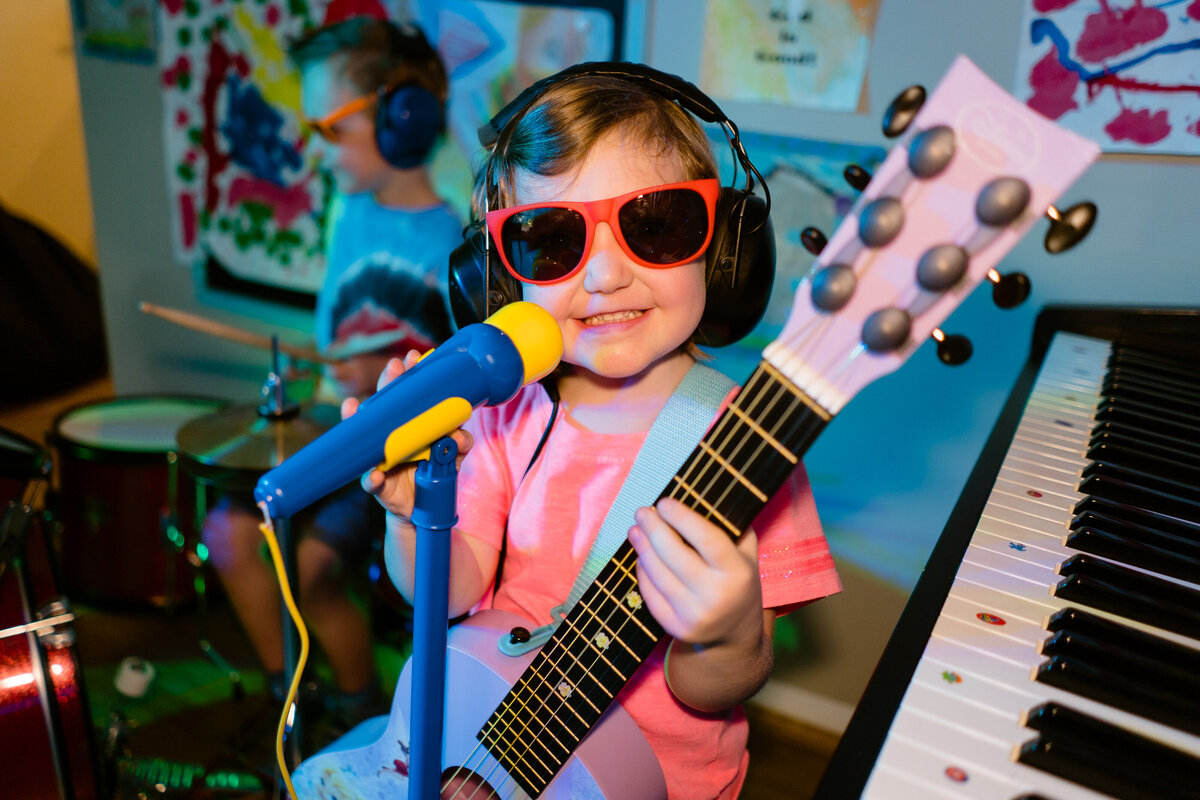 A young child with headphones on holding a guitar and standing in front of a microphone.