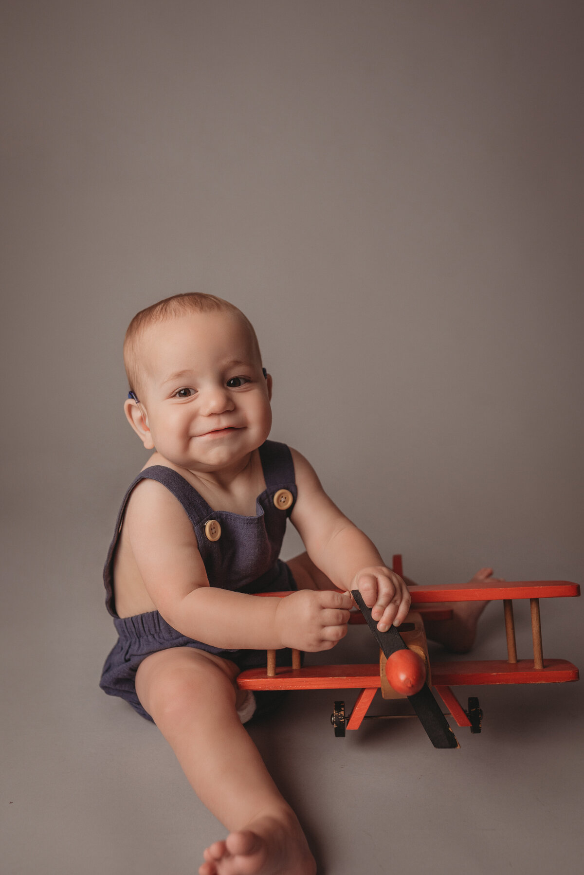 One year old baby boy sitting next to a wooden toy airplane smiling, wearing a dark blue jumper on light gray backdrop