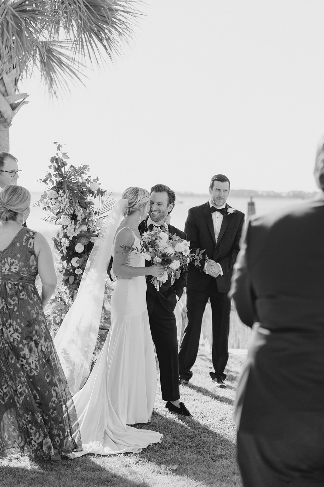Waterfront wedding ceremony at Charleston fall wedding. Bride and groom smile after the first kiss. Palm tree and sunshine. Black and white wedding photos. Kailee DiMeglio Photography.