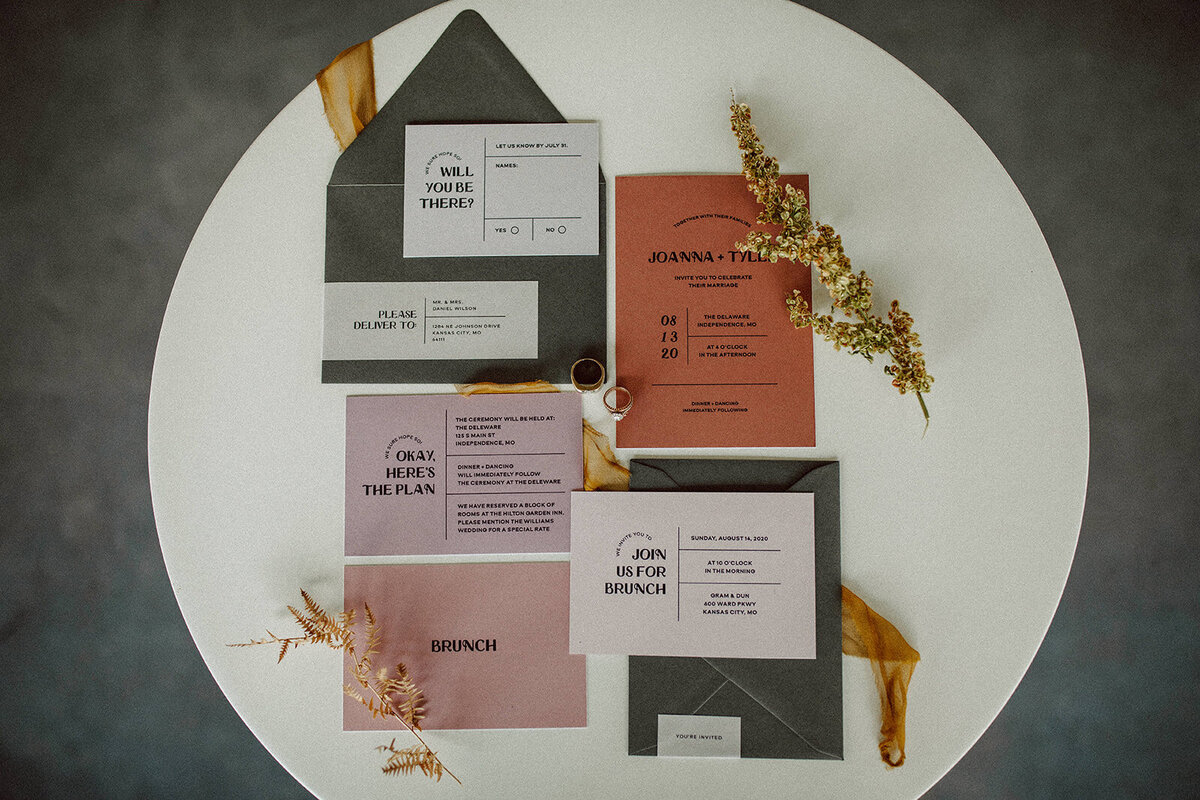 Off-white, peach and mauve-colored wedding stationery with black font and wedding bands atop a white rounded table