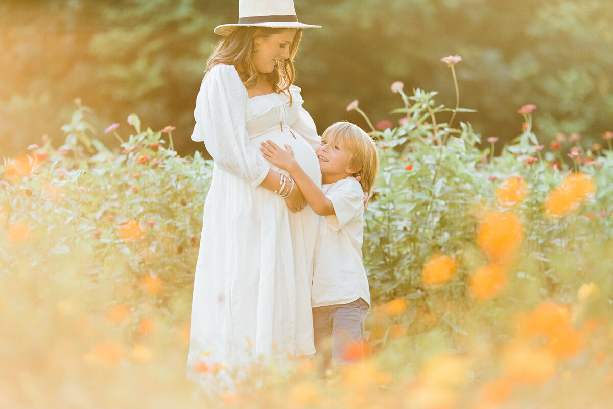 pregnant mom in hat hugs son while standing in a flower garden of orange and multi color flowers.