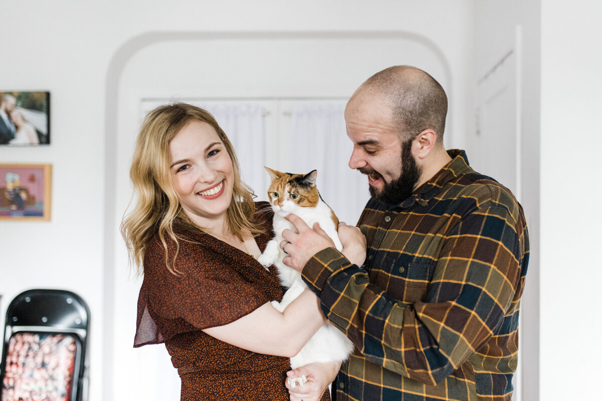 A couple posing playfully with their cat in their apartment during their engagement session in Park Slope in Brooklyn, New York. The woman on the left is wearing a dark orange dress while the man on the right is wearing a dark checkered dress shirt. Their cat is a calico with a white, orange, and black pattern.