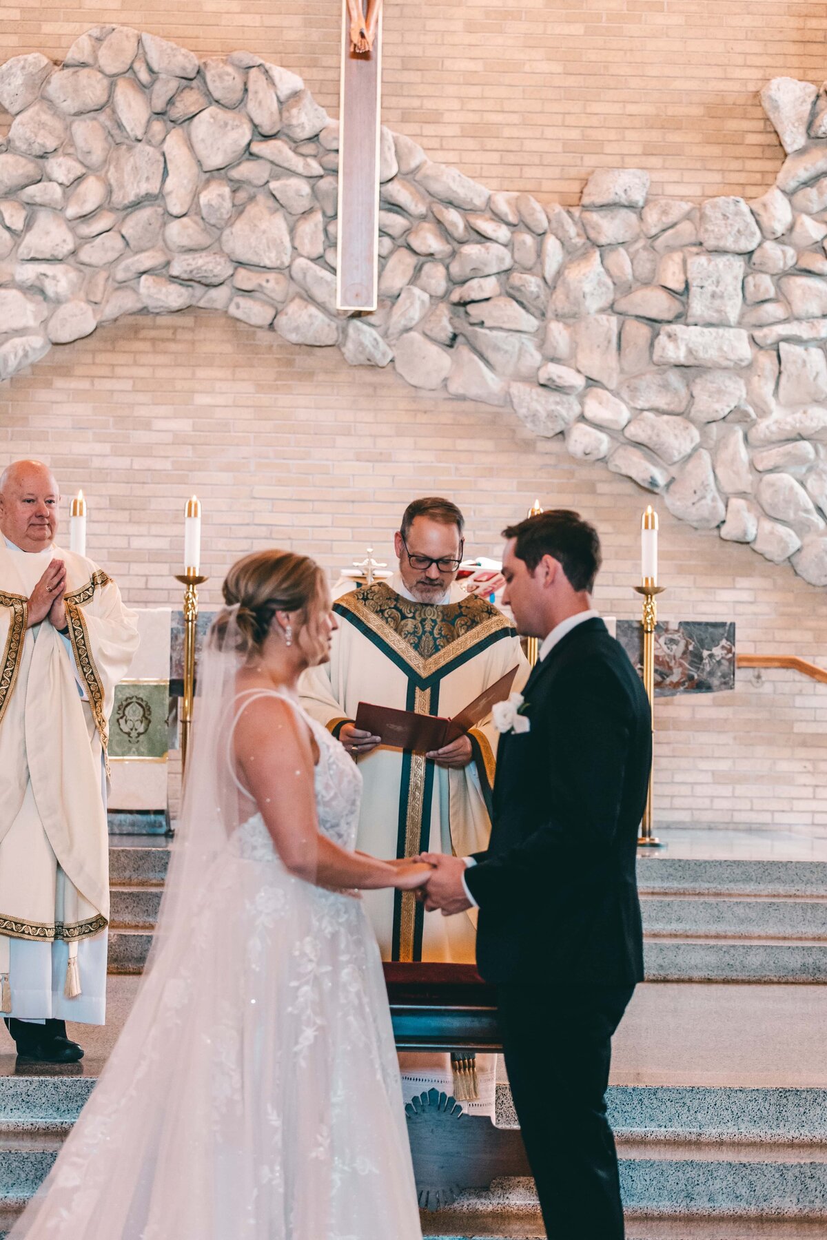 A bride and groom exchange vows in front of a priest at a church altar, with lit candles and a stone backdrop, under the guidance of a wedding coordinator from Iowa.