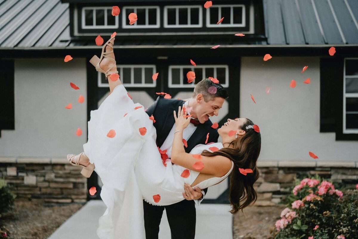 A bride and groom, enveloped in a shower of rose petals, joyfully embrace in front of a house