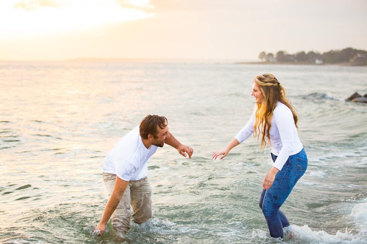 Man splashes his partner as they play at the beach during their Harkness engagement session.
