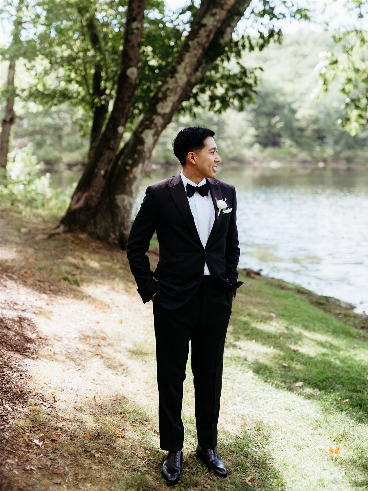 Dapper groom in black suit and bowtie poses under a tree next to a lake at Cedar Lakes Estate in Hudson Valley