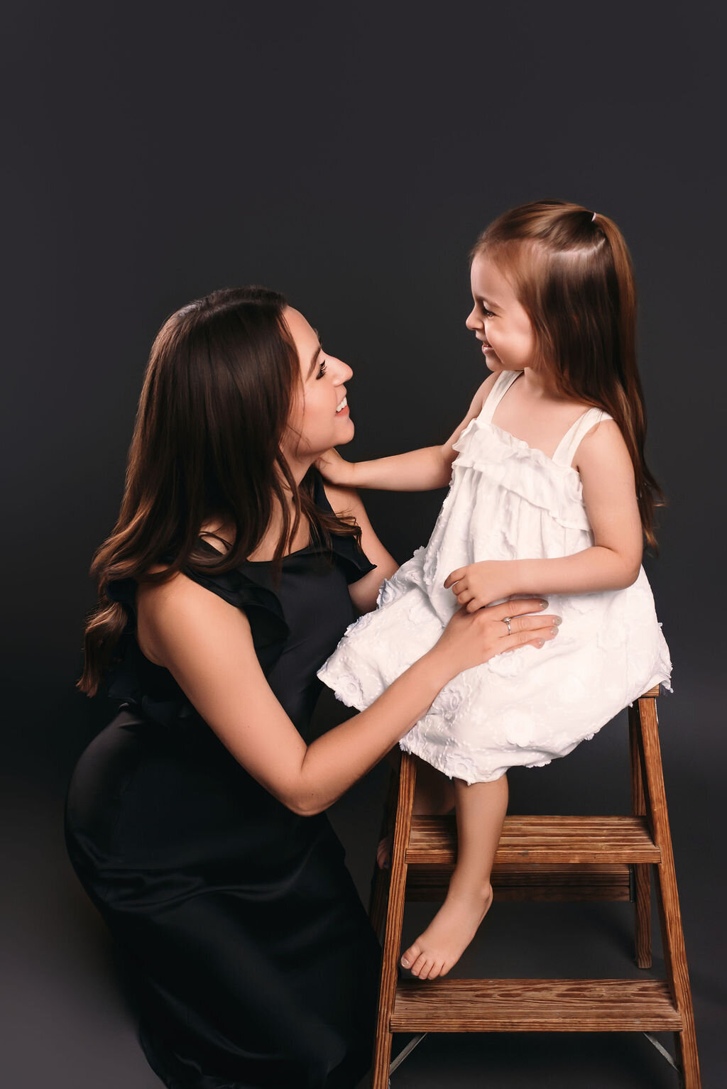 A mother in a black dress and heels sits on the floor in front of a gray-toned backdrop holding her young barefoot daughter in a white dress on a vintage ladder, captured by a photographer in Bemidji, Minnesota.