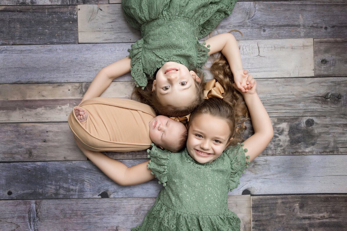 Two young girls in green dresses lay on a wooden floor, forming a circle while smiling up at the camera. Between them, a sleeping baby is wrapped in a beige swaddle. The girls' heads rest close to the baby's. This precious moment was beautifully captured by a Jacksonville baby photographer.