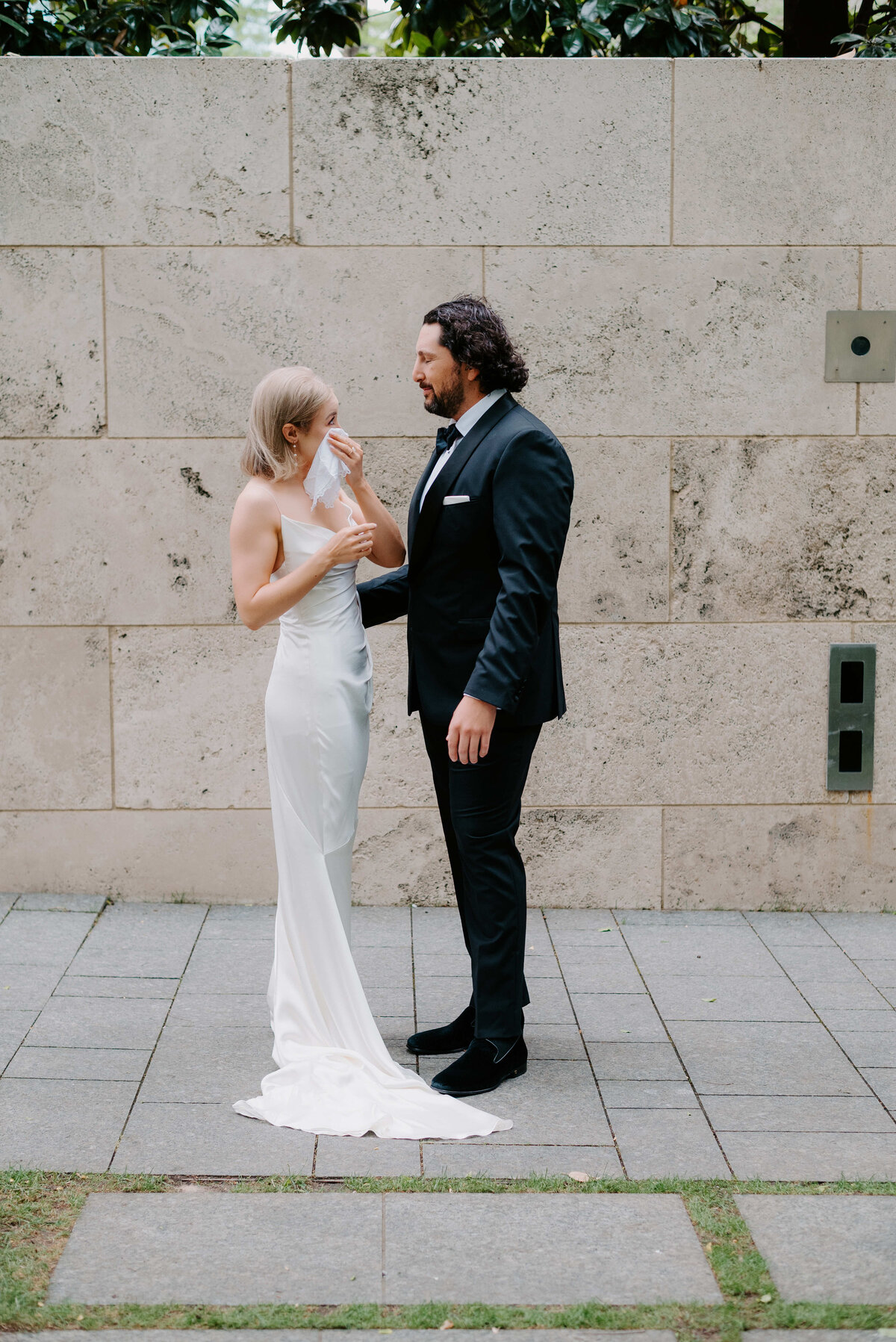 A bride in a white dress wipes tears while a groom in a black tuxedo comforts her. They stand on a stone path with a textured wall background, evoking a heartfelt moment.