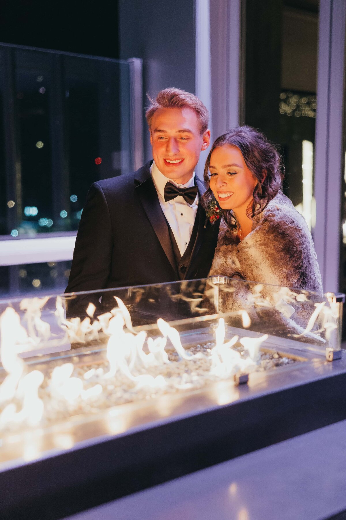 A couple in formal attire smiling and looking at a fire pit with city lights in the background, enjoying an event in Davenport at night.