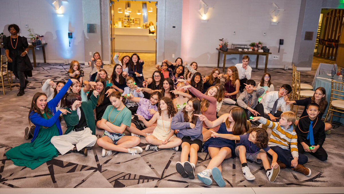 A large group of friends sit on a ballroom floor pointing at the mitzvah girl in the center