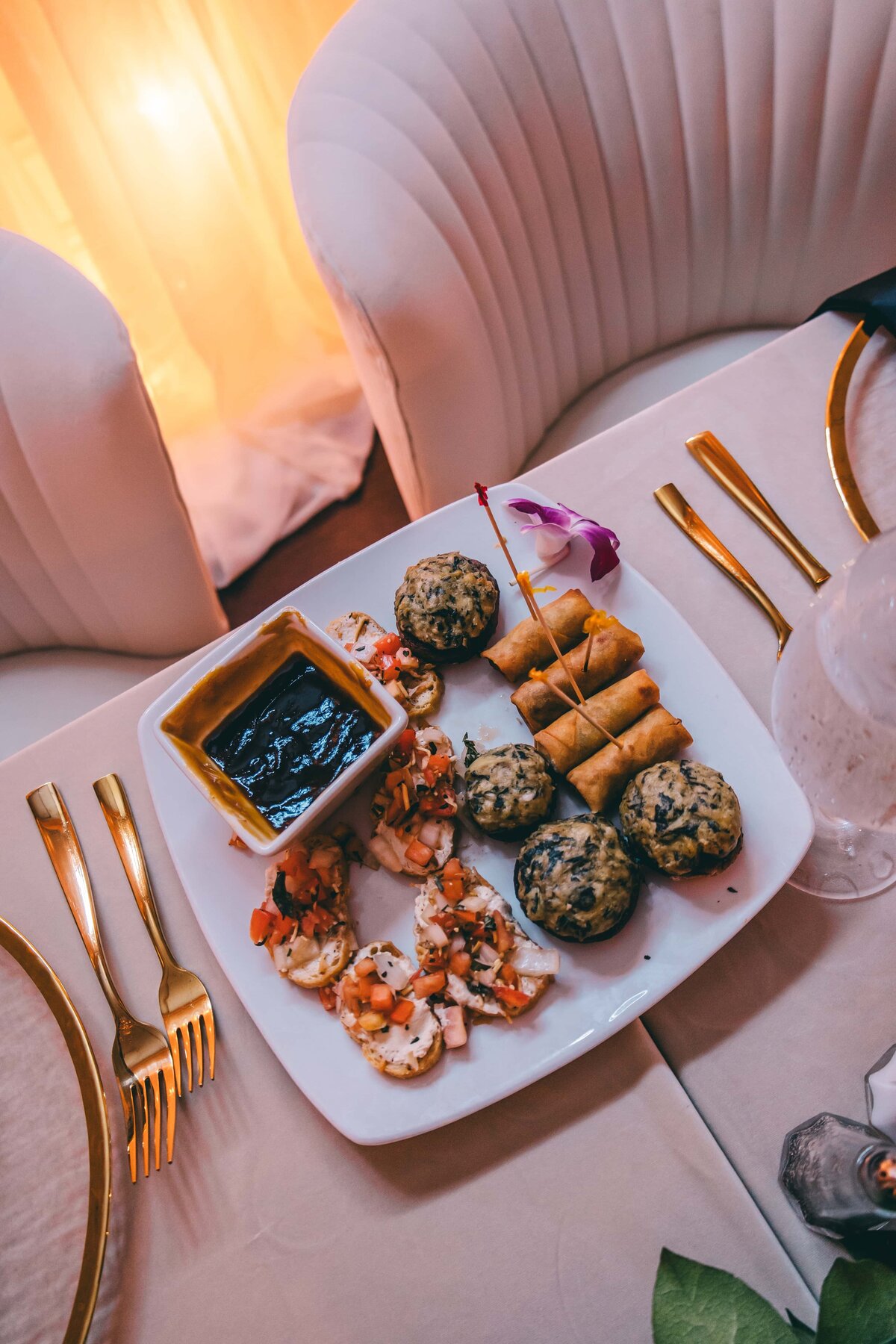 A plate of assorted appetizers including spring rolls, meatballs, and bruschetta, on a table with elegant gold cutlery at a wedding coordinator's showcase in Iowa, against a soft-l