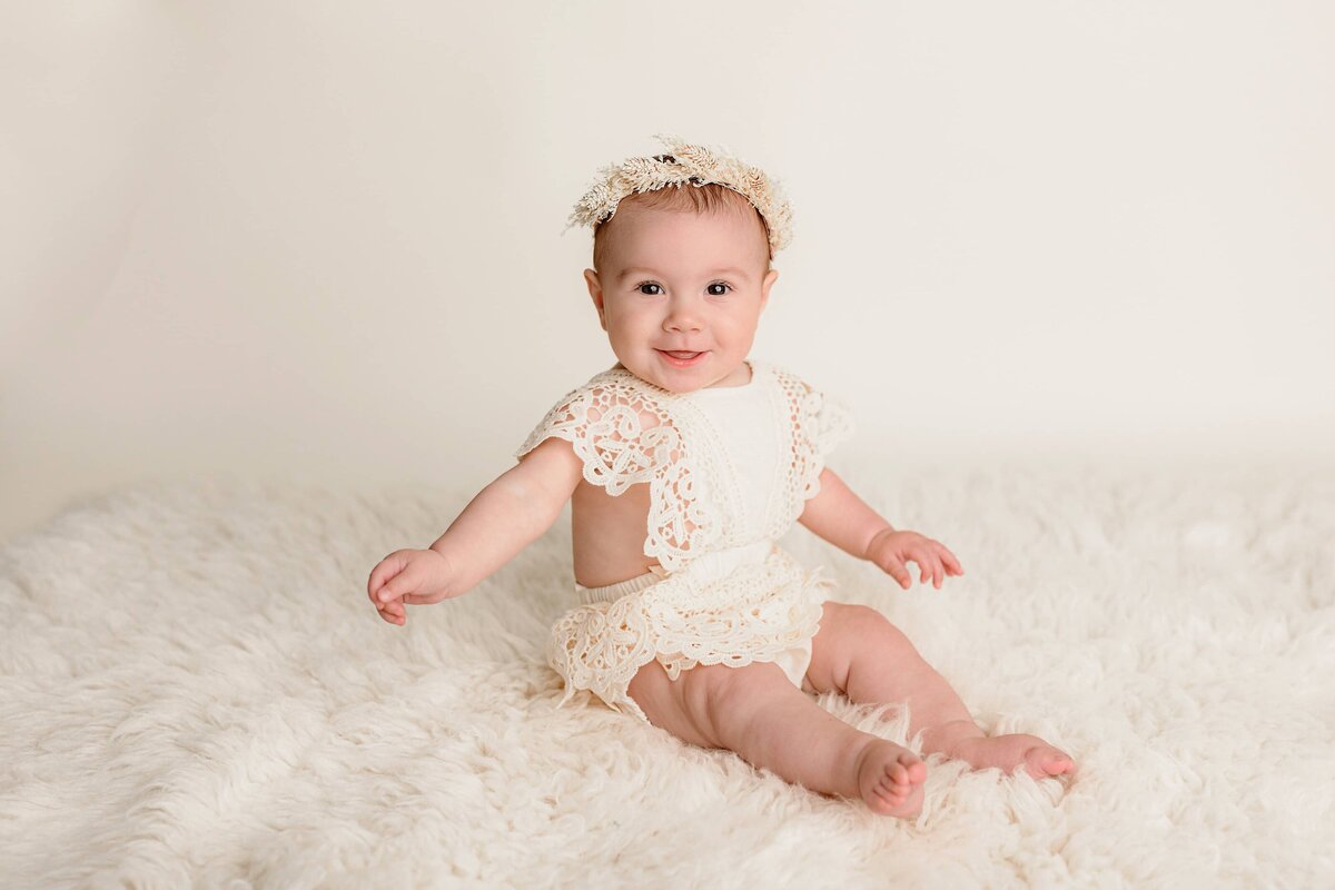 7-month-old little girl sits in a boho style Romper with a headband sitting on a cream colored rug smiling