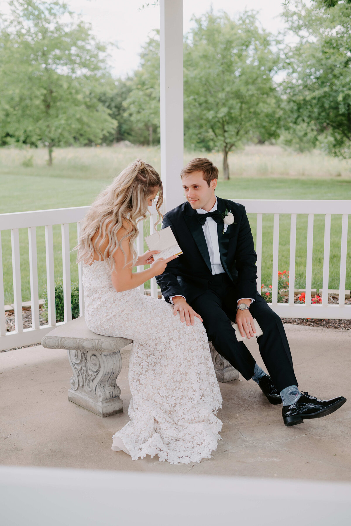 A bride in a lace dress and groom in a black tuxedo sit on a stone bench in a gazebo, exchanging letters. The scene is serene and intimate, surrounded by greenery.