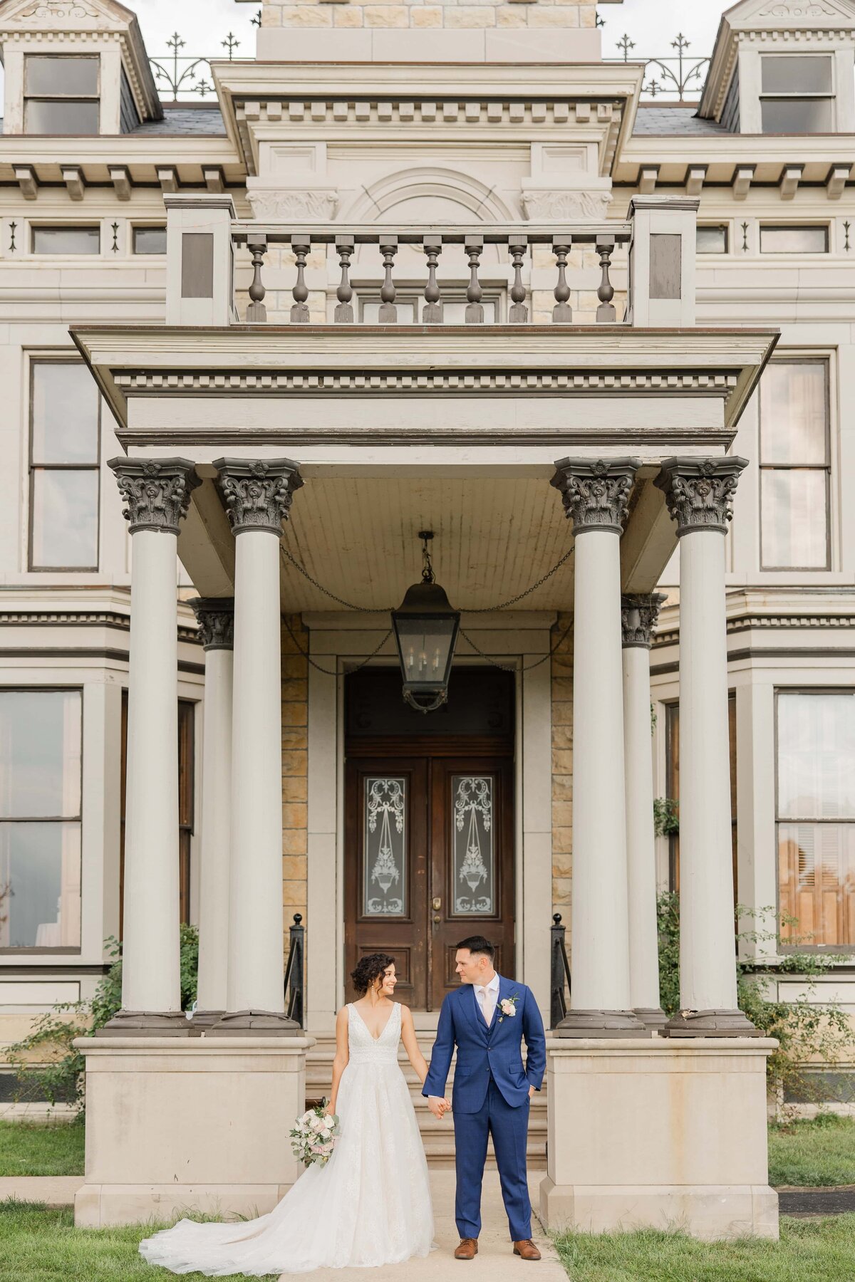 A bride in a white gown and a groom in a blue suit holding hands in front of an ornate historical mansion with columns and balconies at an event in Davenport.