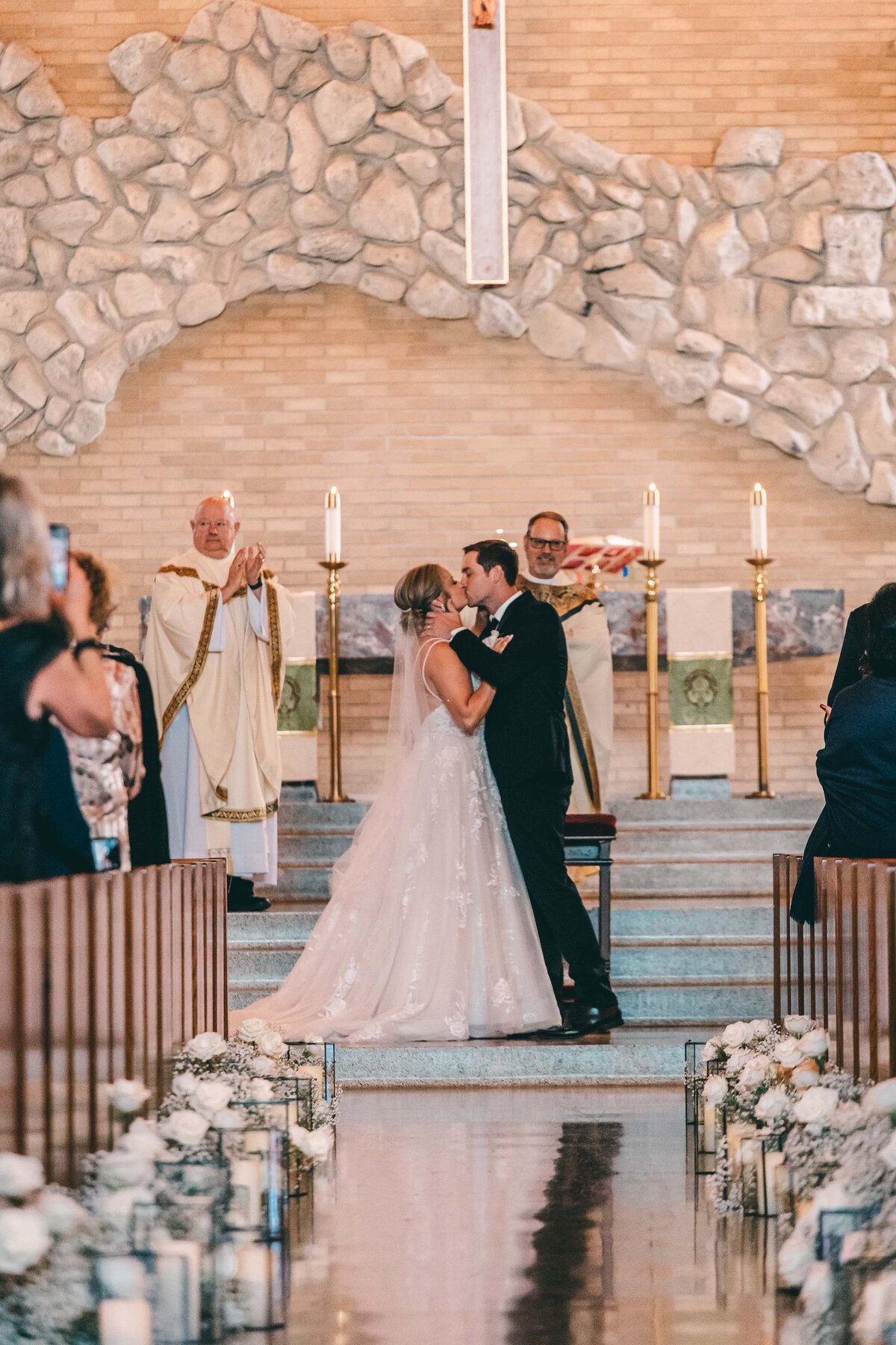 A bride and groom kissing at the altar of a church in Iowa, with a priest officiating and guests watching. The background features a large stone arch.