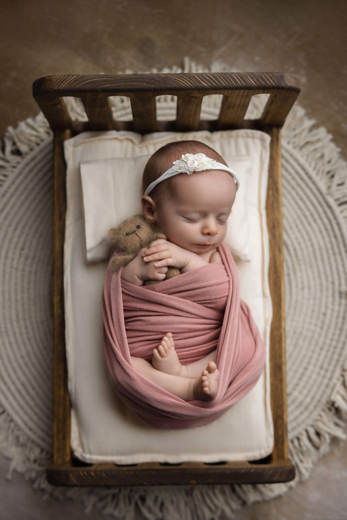 newborn baby wrapped in pink fabric holding a tiny teddy bear sleeping in a tiny wooden bed