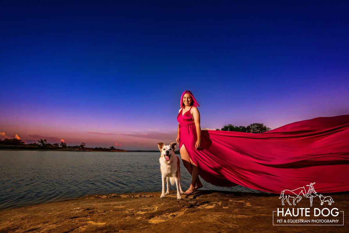 Medium sized white and brown dog smiles at the camera next to a woman on the shore of Grapevine Lake. The woman is wearing a long pink flying dress fluttering at sunset.