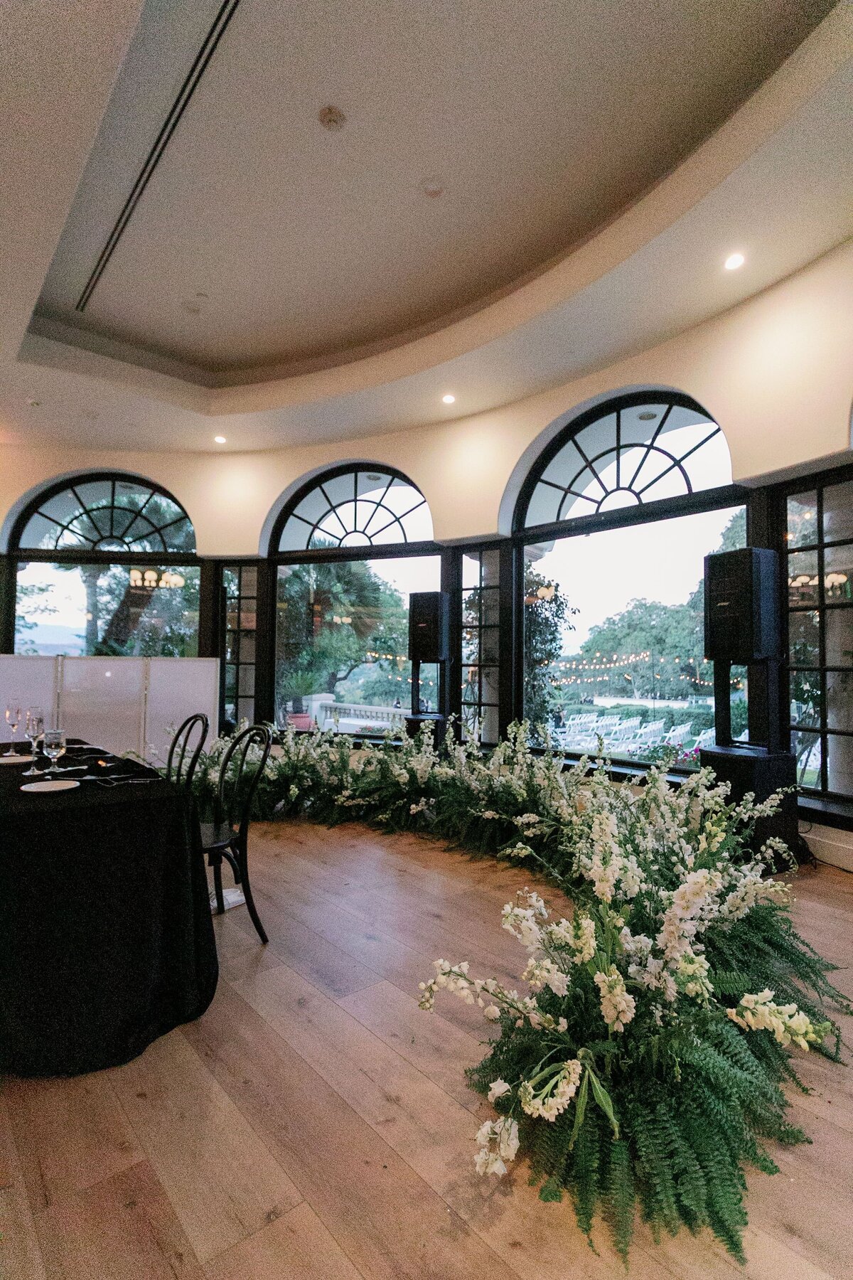A half moon full of white flowers and greenery lines the floor behind the bridal dining table.