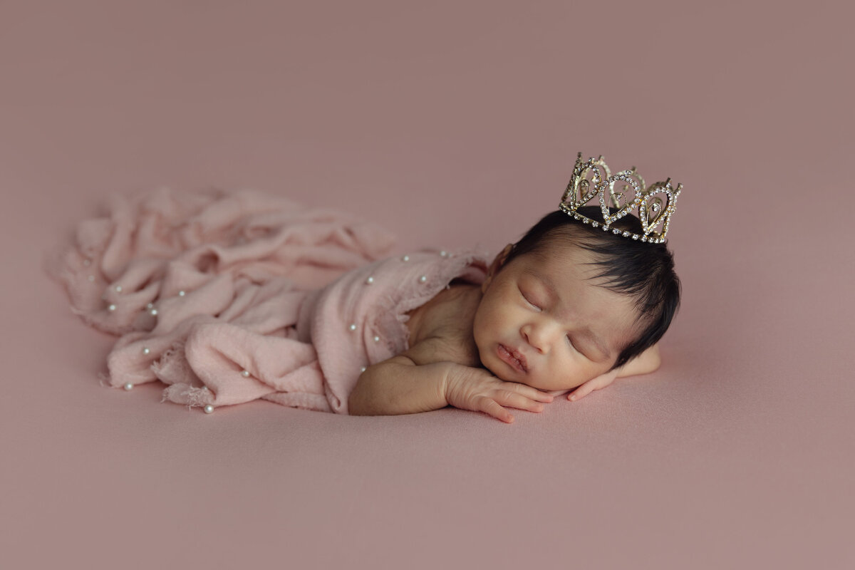 A newborn baby girl sleeps under a pink blanket wearing a tiny crown in a NJ Newborn Photography studio
