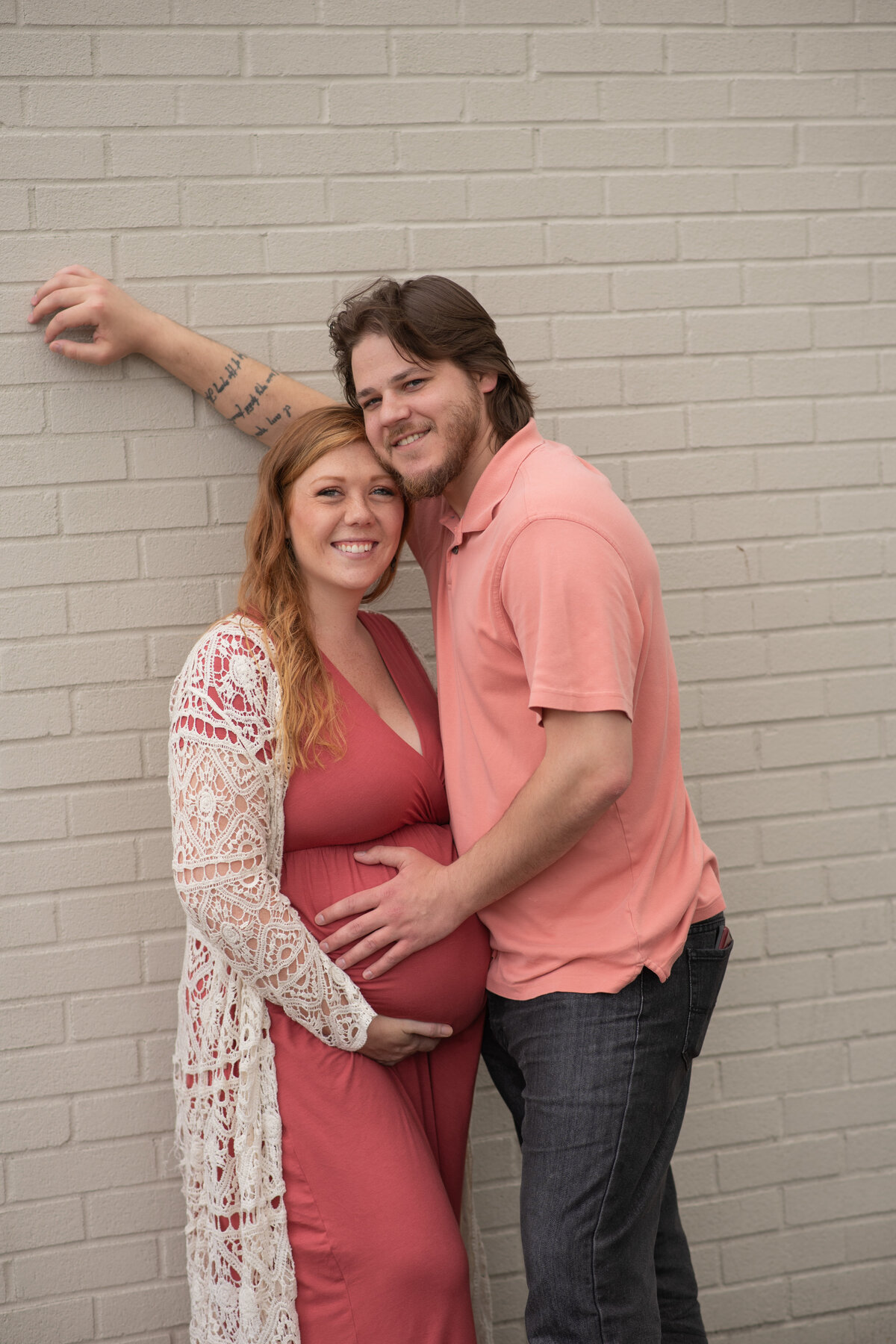 Husband & Pregnant wife snuggling and leaning on white brick wall
