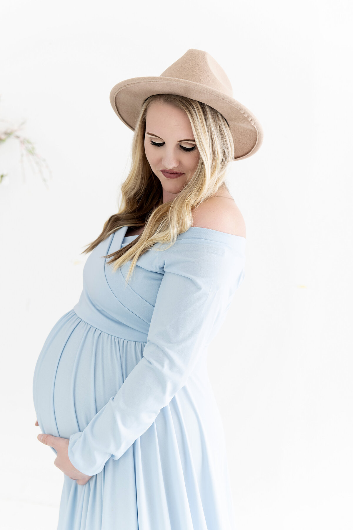 A pregnant woman in a flat brimmed hat and blue maternity gown happily holds her bump while standing in a studio