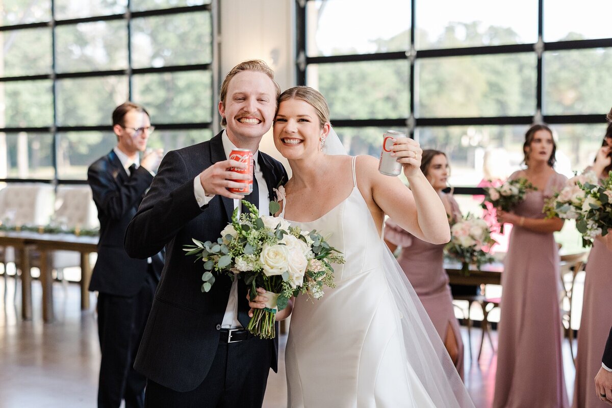 A bride and groom posing for a quick photo during their wedding day in DFW, Texas. They're both smiling and holding up cans of soda in a celebratory toast. The groom is on the left and is wearing a dark suit and boutonniere. The bride is on the right and is wearing a sleeveless, white dress and veil, and is holding a bouquet. Multiple members of the wedding part can be seen in the background.