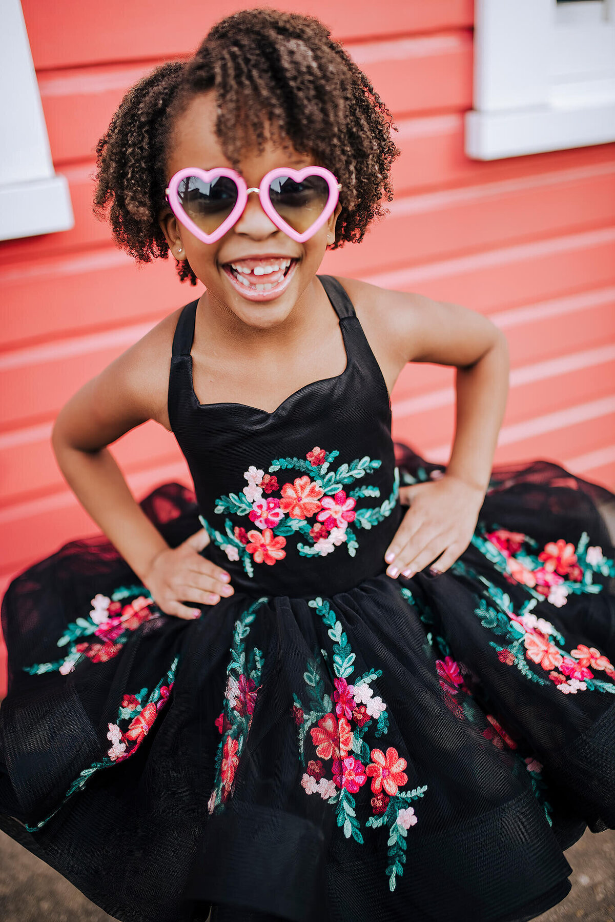 Young African American girl wearing pink hearts glasses laughing and smiling in a black floral dress at Fells Point in Baltimore Maryland