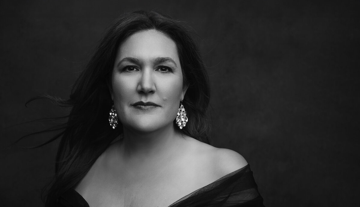 Timeless Cincinnati portrait of a confident woman draped in an off-shoulder ensemble, accentuated by dazzling earrings. The monochrome tone highlights her strong features against a soft black background.