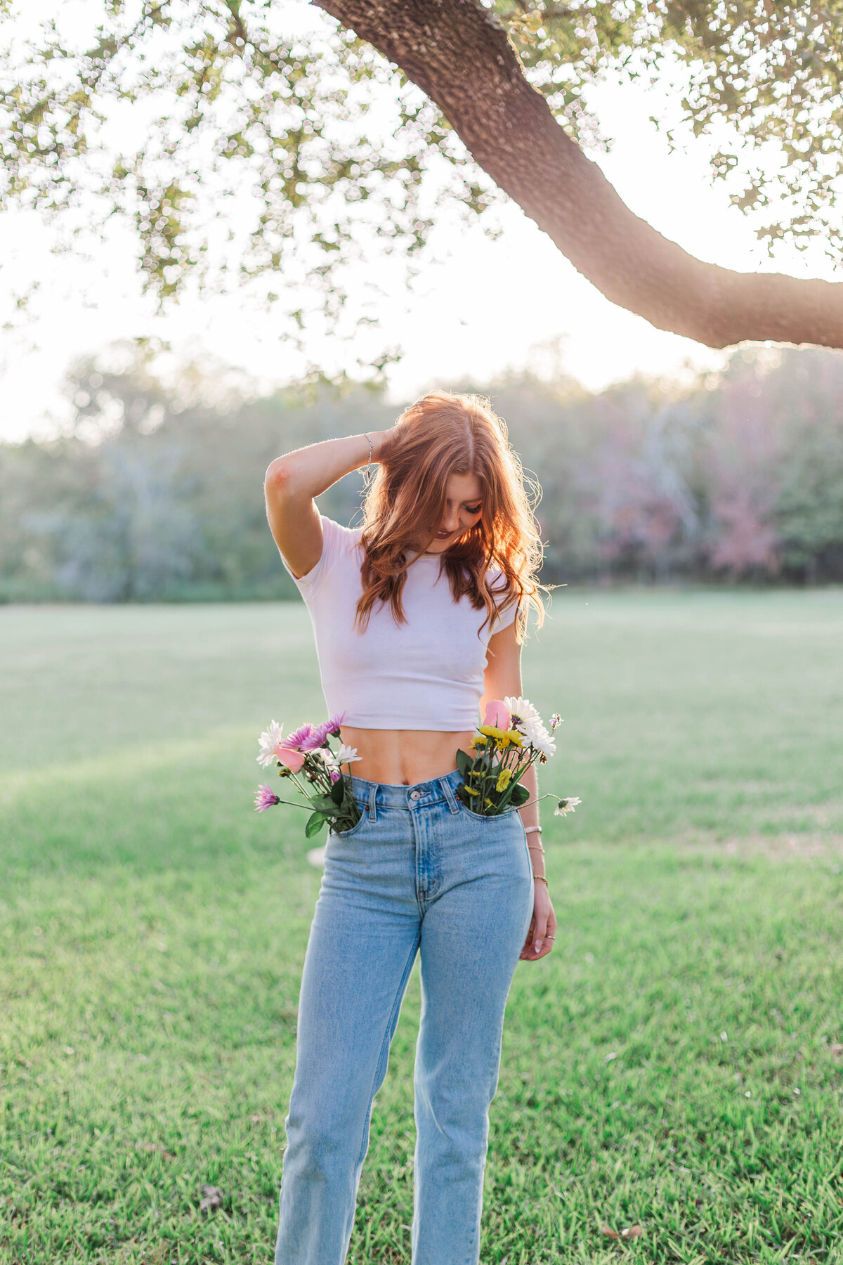 High school senior looks down at her feet with her hand in her hair while standing in a field with flowers coming out of her pockets