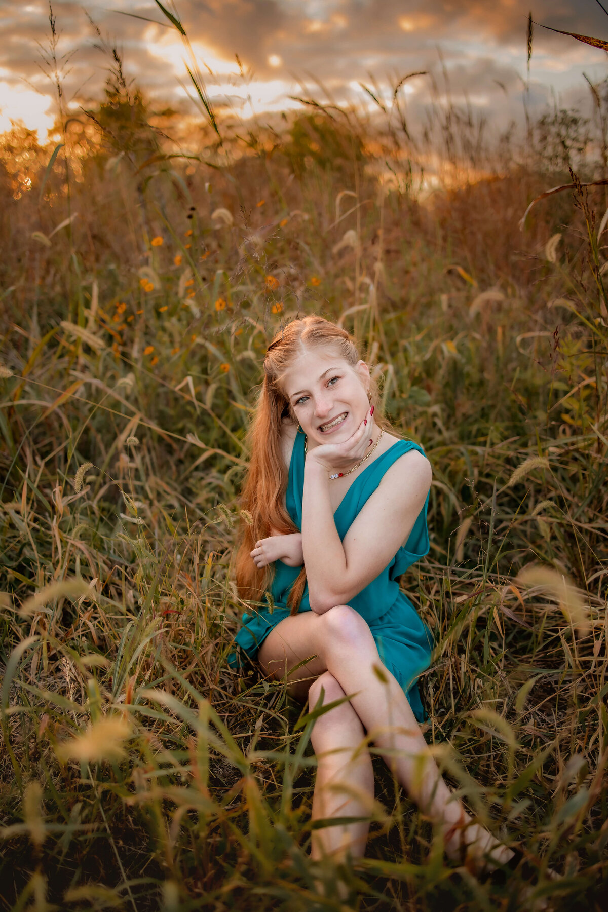 A blonde haired girl is wearing a blue romper for her high school senior photos and sitting in a field with a beautiful sky.