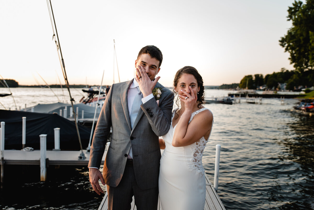 bride and groom stand on dock at sunset and hold their ring hands up by face