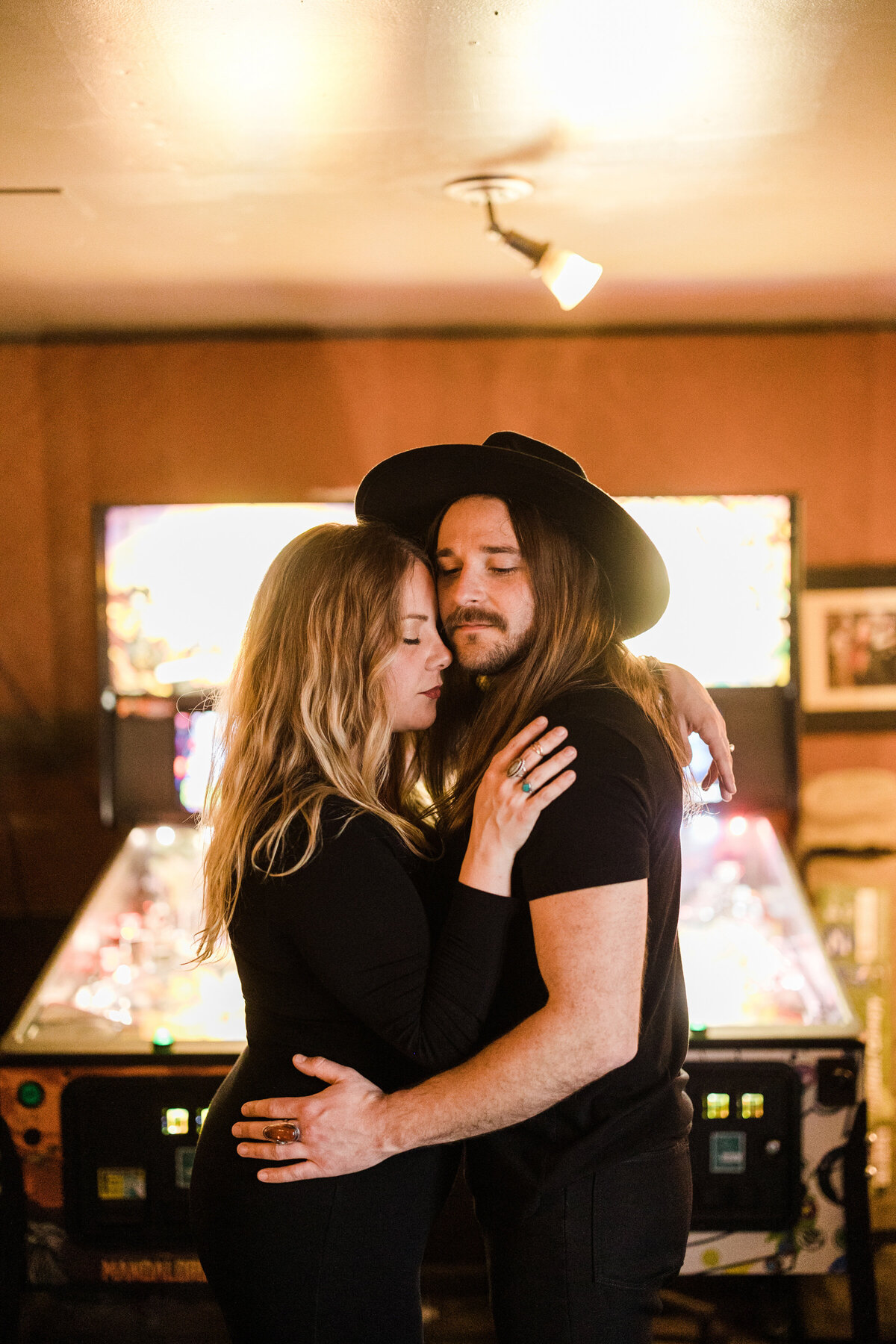 A couple holding each other close with closed eyes during their engagement photoshoot in Fort Worth, Texas. Both are dressed in all black and the man on the right has a black, large brimmed hat on. They both stand in front of multiple pinball machines.