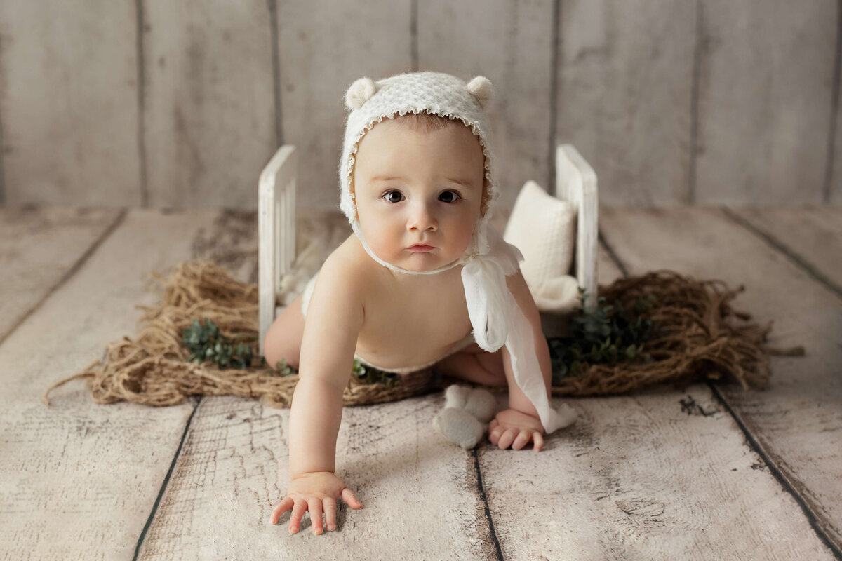 toddler in with  a bear bonnet crawling       in  front of       a        wooden bed at a toddler photography photo session            a  photography photo session laying on a white backdrop with a bow in her hair in a northern virginia stuio