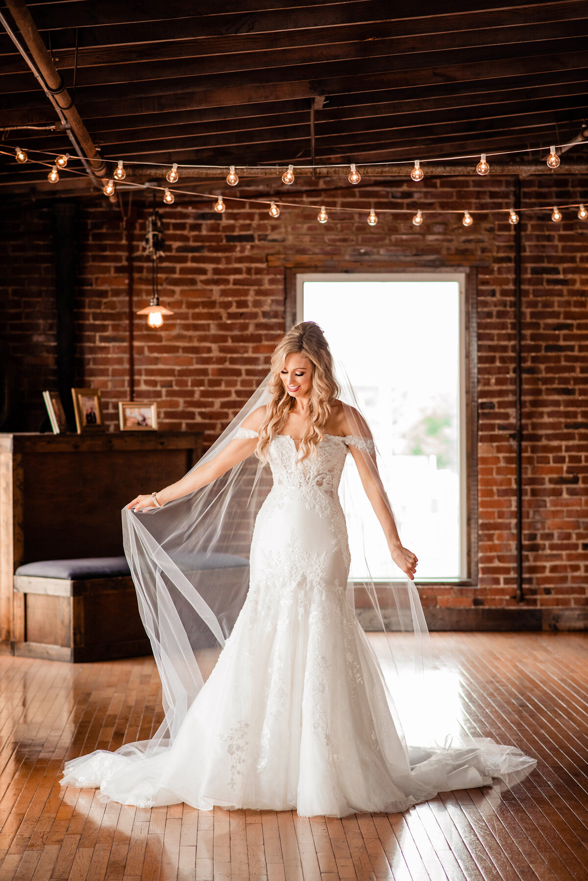 Bride playing with her cathedral veil inside wedding venue with brick walls and string lights