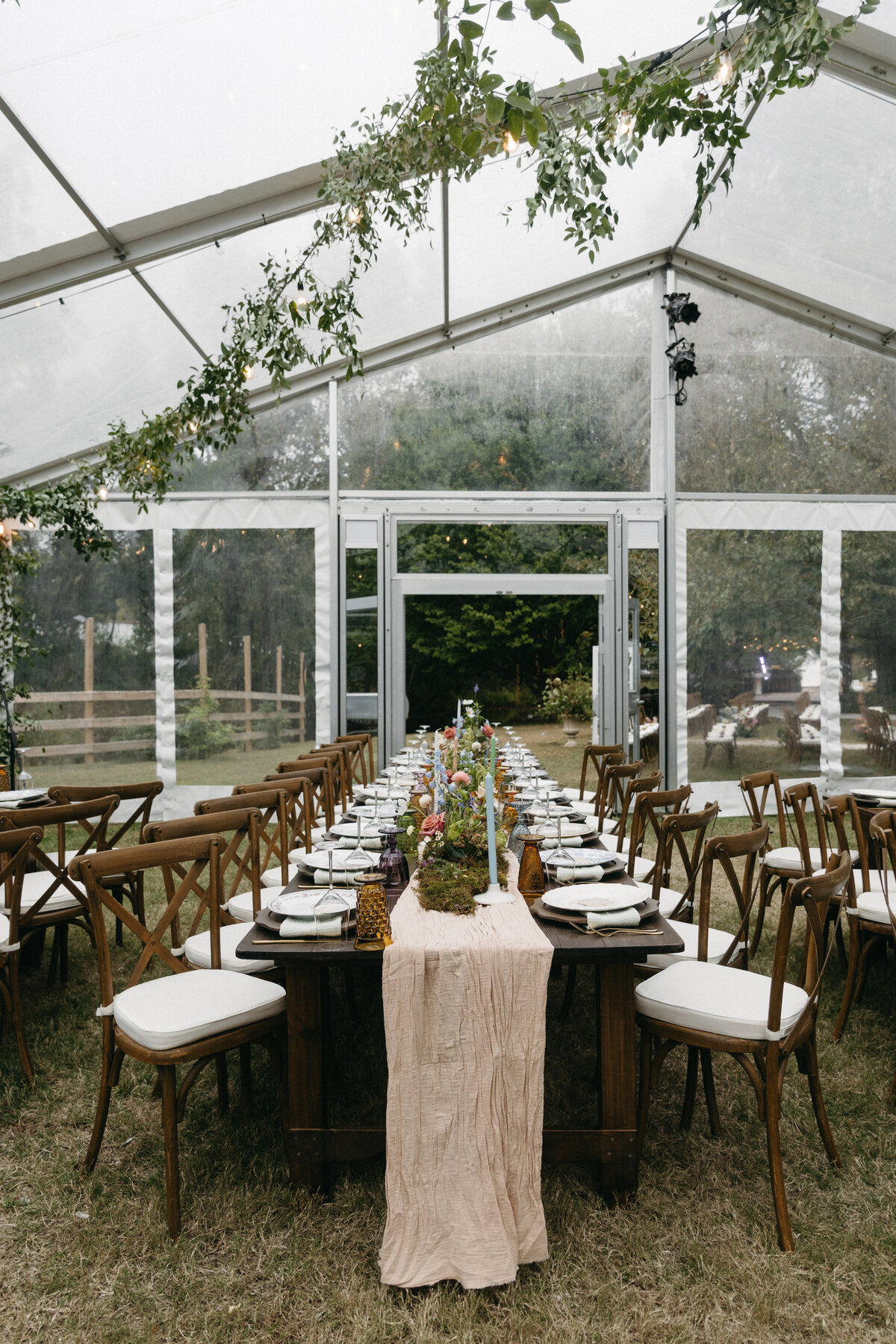 Long family-style wedding table set inside a clear-top tent with romantic greenery installations, pastel florals, layered table settings, and warm candlelight for an elegant garden celebration.