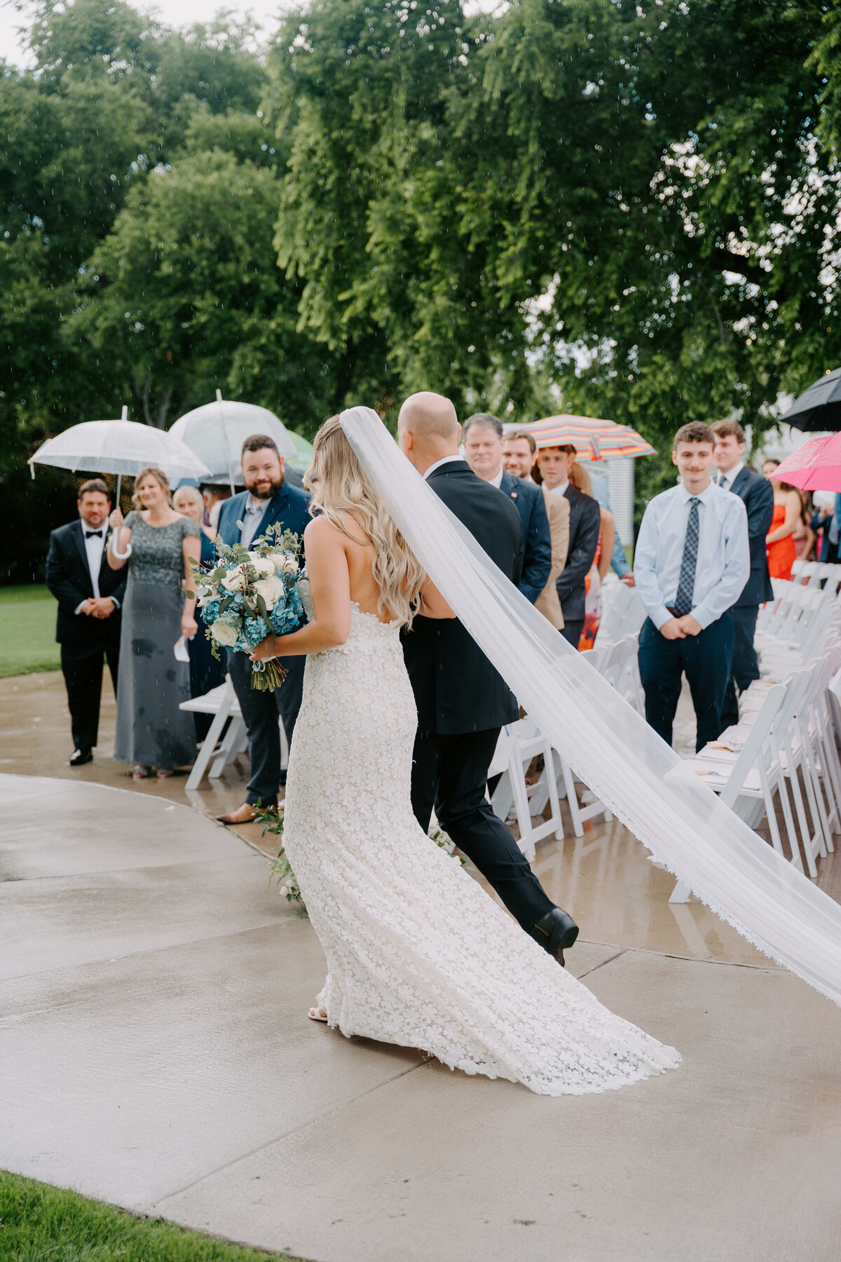 Bride in a lace gown with a long veil holds a bouquet, walking down the aisle outdoors. Guests with umbrellas line the sides, creating a joyful atmosphere.