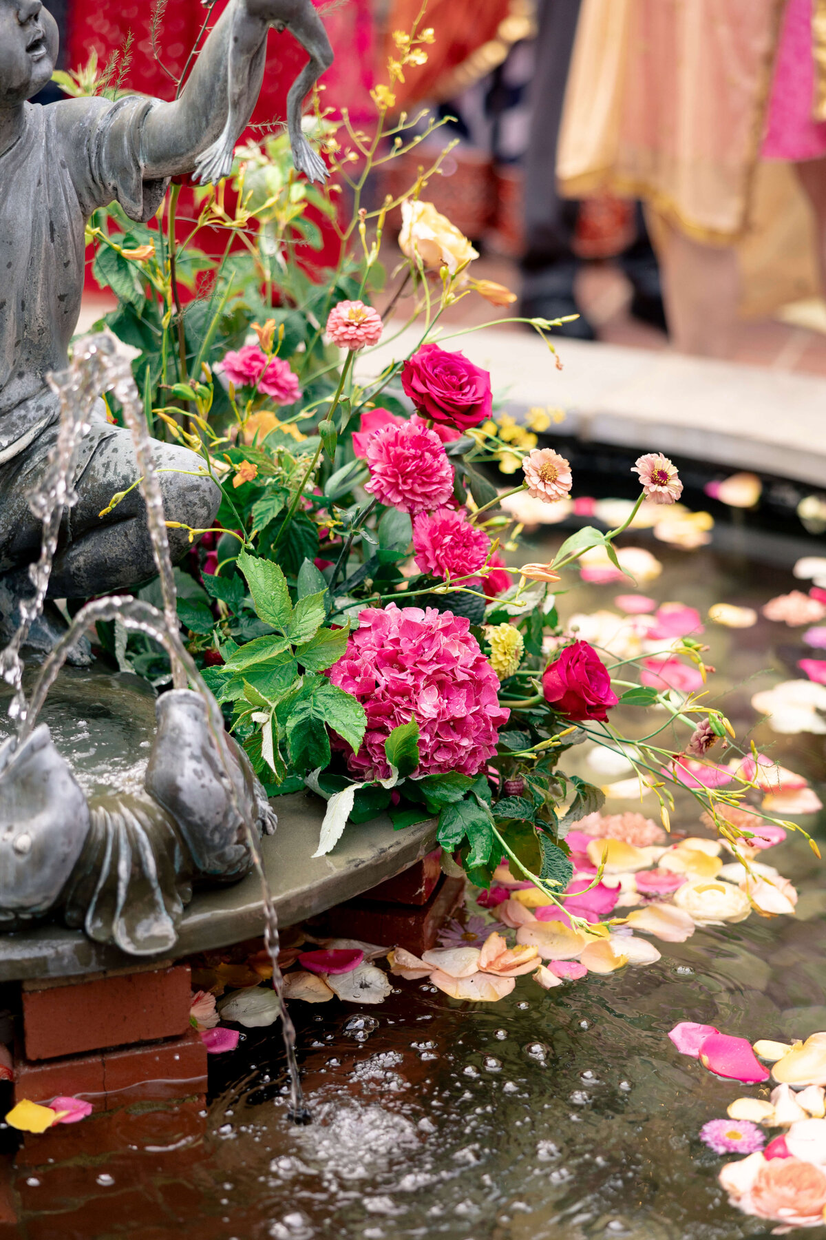 A stone fountain with flowing water features a statue sitting on the edge. Nearby, vibrant pink and red flowers with green leaves are arranged artistically. Colorful flower petals are scattered on the surface of the water.