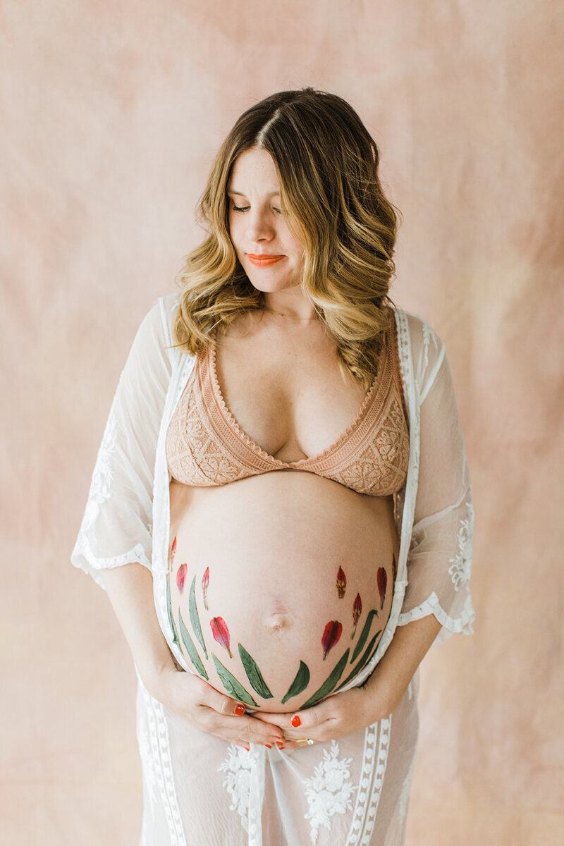 pregnant mom stands with flower petals adhered to bare belly in front of a blush backdrop