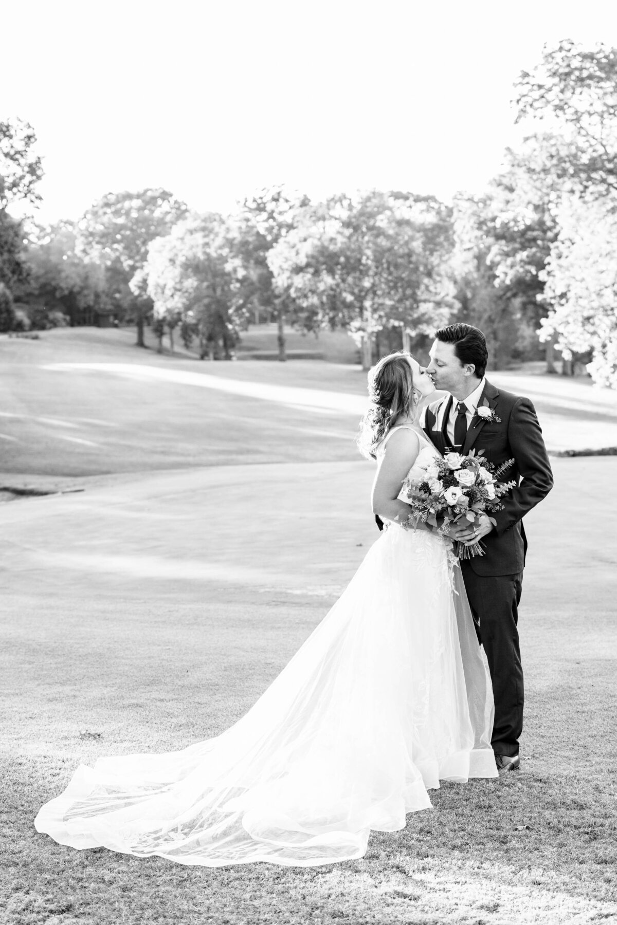 Black-and-white-image-of-bride-and-groom-kissing-while-holding-the-bride's-bouquet-with-the-rolling-hills-of-Pine-Island-Country-Club's-golf-course-in-the-background