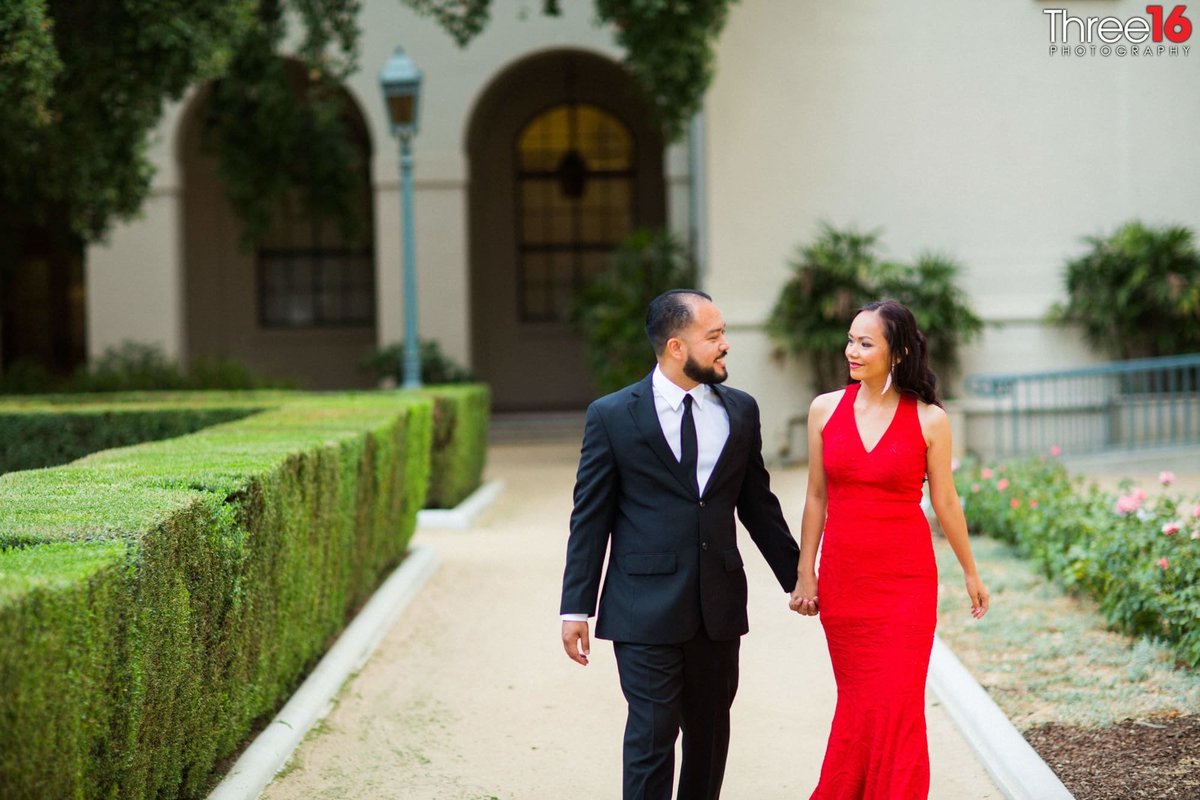 Engaged couple walk along the Pasadena City Hall while holding hands and looking at each other
