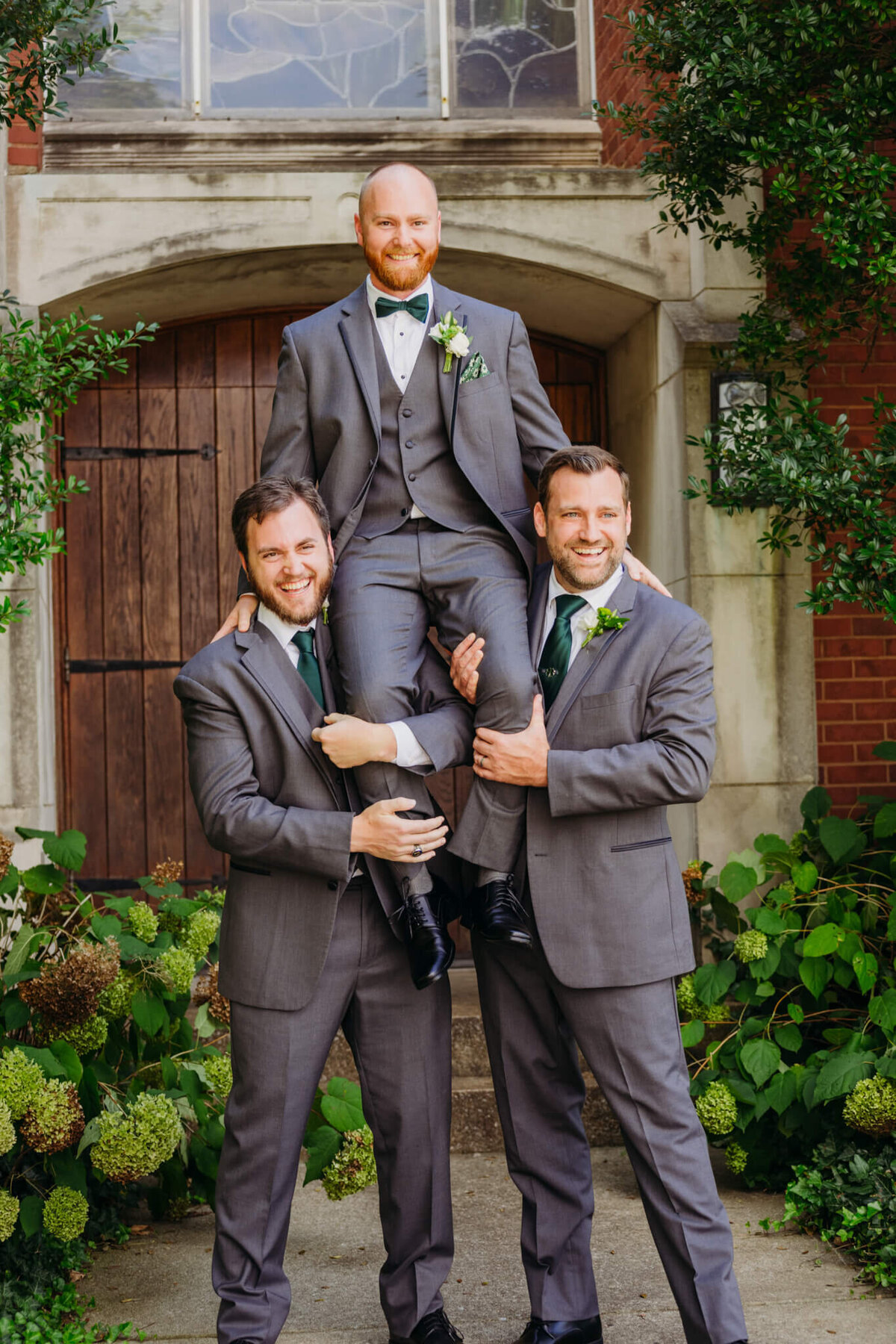 photo of a Groom on two groomsmen shoulders while everybody laughs in front of a wooden door and hydrangea bushes