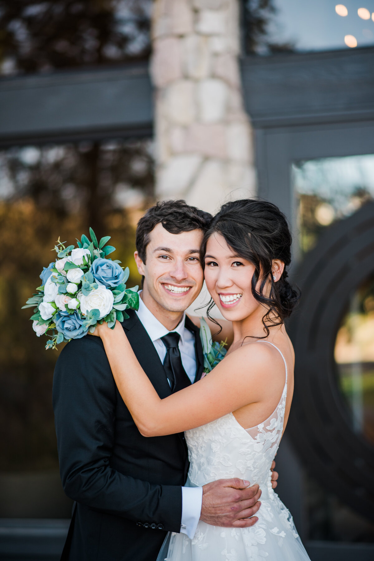 denver wedding photographer captures groom embracing his bride around the waist as she holds his shoulder with her blue and green wedding flowers in one hand at their outdoor colorado wedding