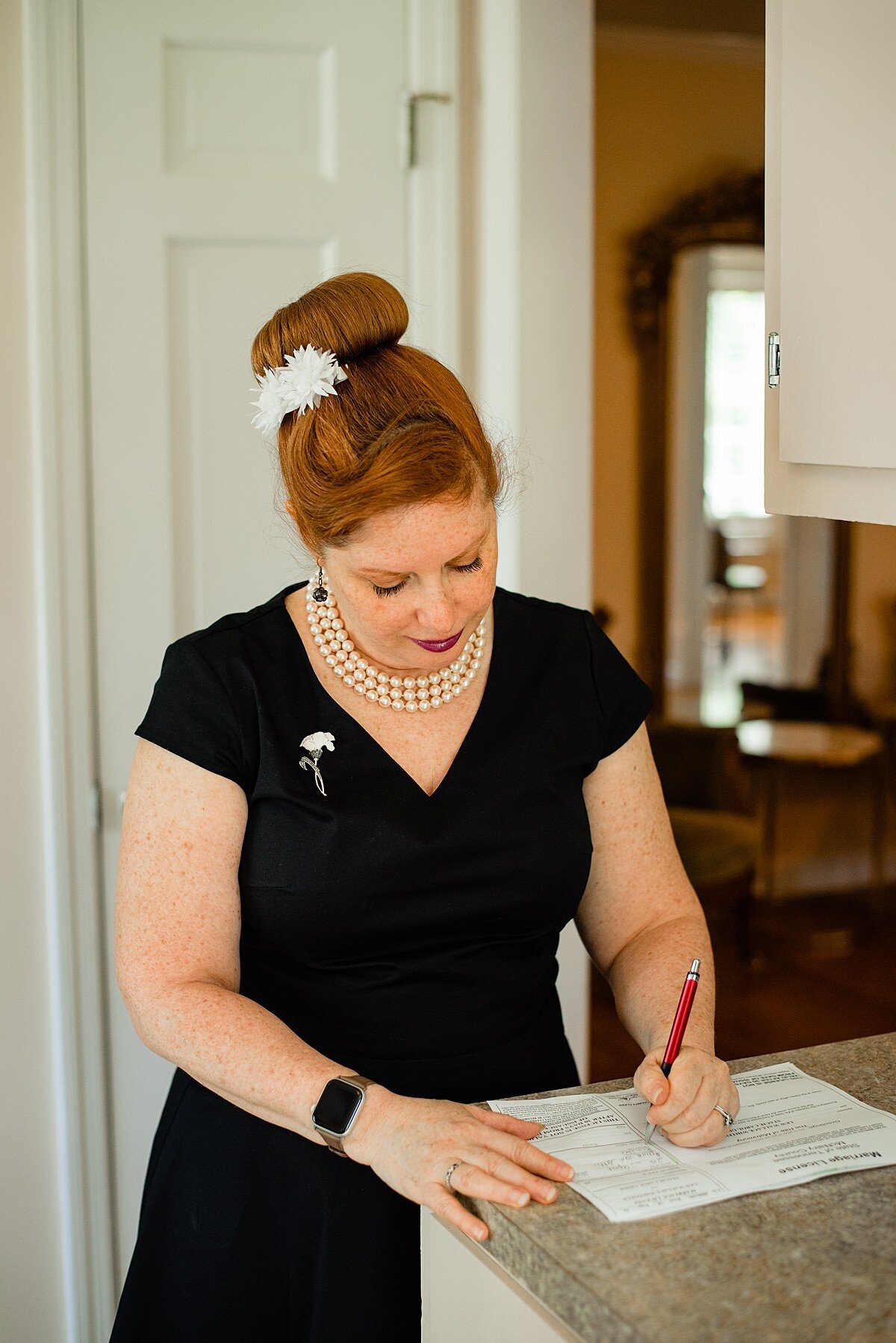 Raina, the wedding officiant signs the marriage license. The officiant is wearing a black dress with a v-neckline a three strand pearl necklace and a white and obsidian flower pin. She has her red hair swept up into a structured bun and small white flowers as accents.