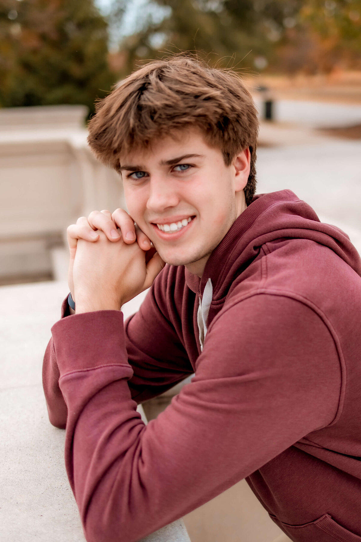 A young man is posed with his hands  closed together next to his face and elbows resting on a concrete ledge  at the Grand Basin for his Senior photos.