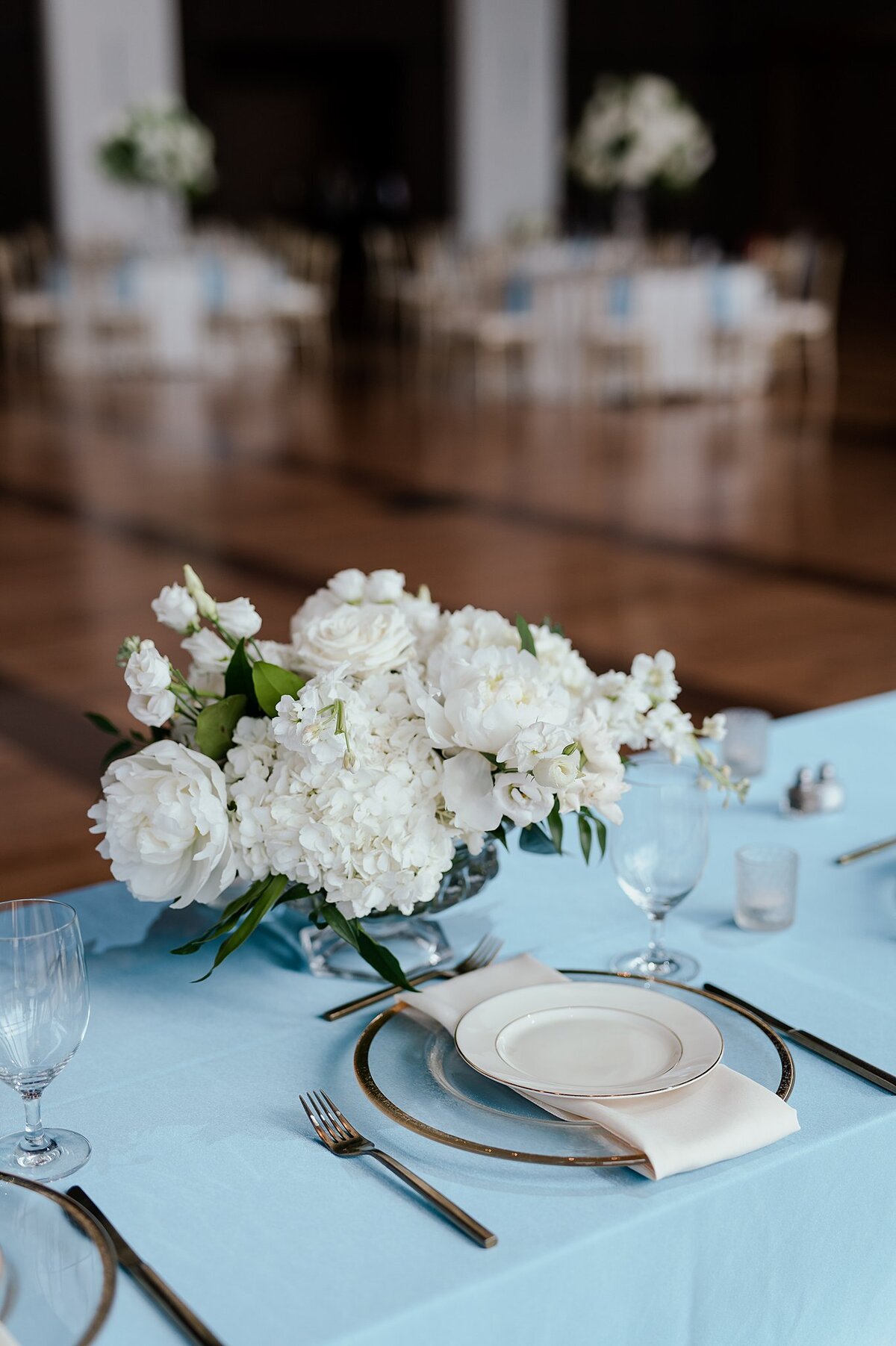 light blue sweetheart table with a low white floral centerpiece. The table at the Country Music Hall of Fame is set with a glass charger with a gold band, a white salad plate with a gold band and gold silverware.